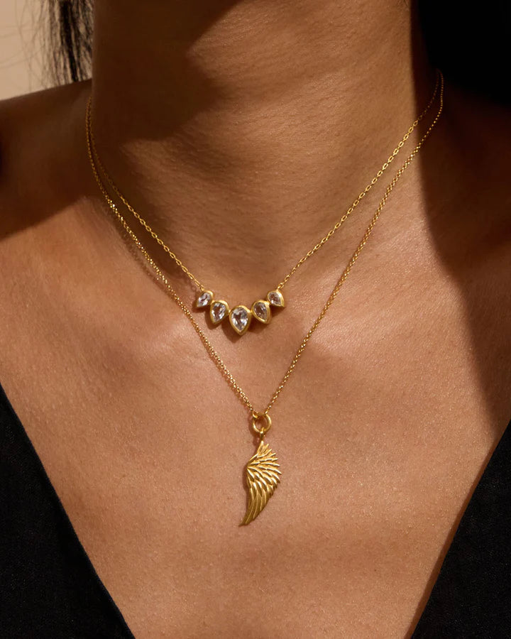 Close-up of a person wearing two gold necklaces with a wing pendant on a neutral background