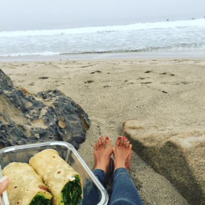 Kimberly sitting at beach with bare feet while eating a healthy wrap. 