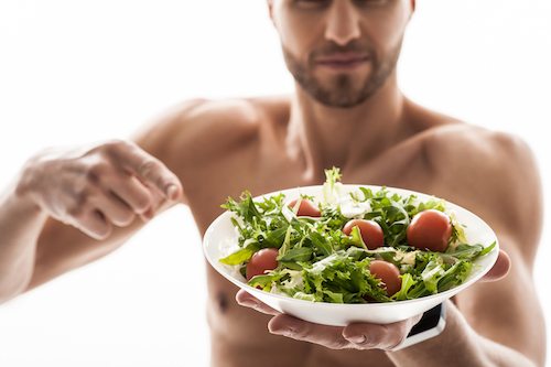 Strong male athlete prefers healthy food. He is standing and pointing finger at salad with proud. Focus on plate. Isolated