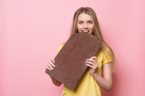 Picture of woman with chocolate smiling. Cute girl holding and eating giant cocoa chocolate bar near pink wall.