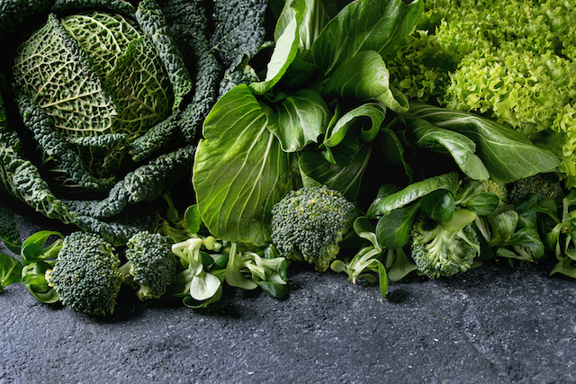 Variety of raw green vegetables salads, lettuce, bok choy, corn, broccoli, savoy cabbage as frame over black stone texture background.