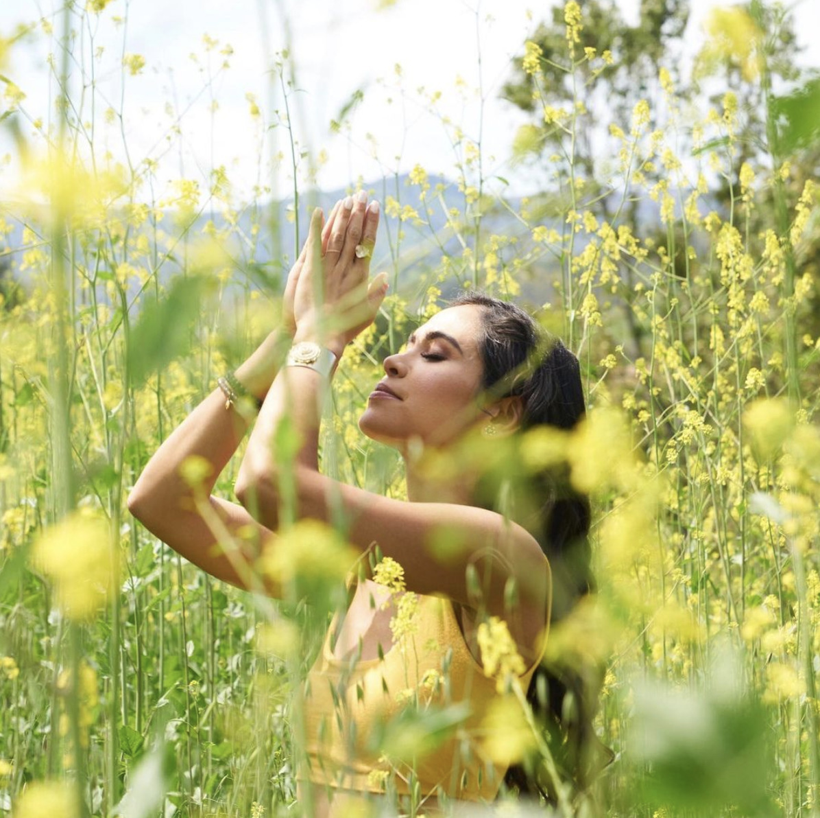 Kimberly Snyder standing in tall grass with palms together and towards the sky in meditation stance. 